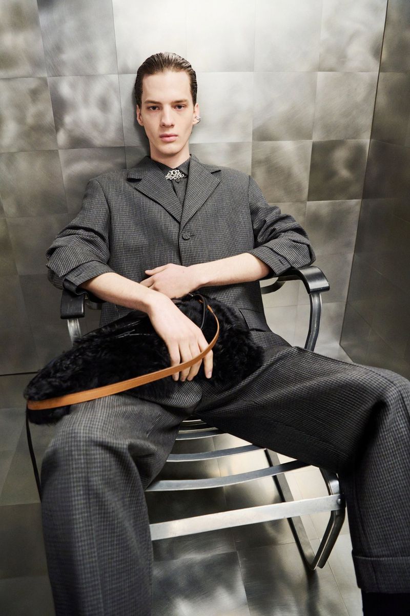 A fair-skinned adult male seated in a metal chair, wearing a gray suit and holding a black fur clutch with a tan strap, indoors.