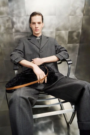 A fair-skinned adult male seated in a metal chair, wearing a gray suit and holding a black fur clutch with a tan strap, indoors.