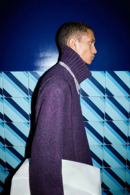 Profile shot of a black male wearing a purple wool sweater with a silver chain necklace. He stands against a blue tiled wall.