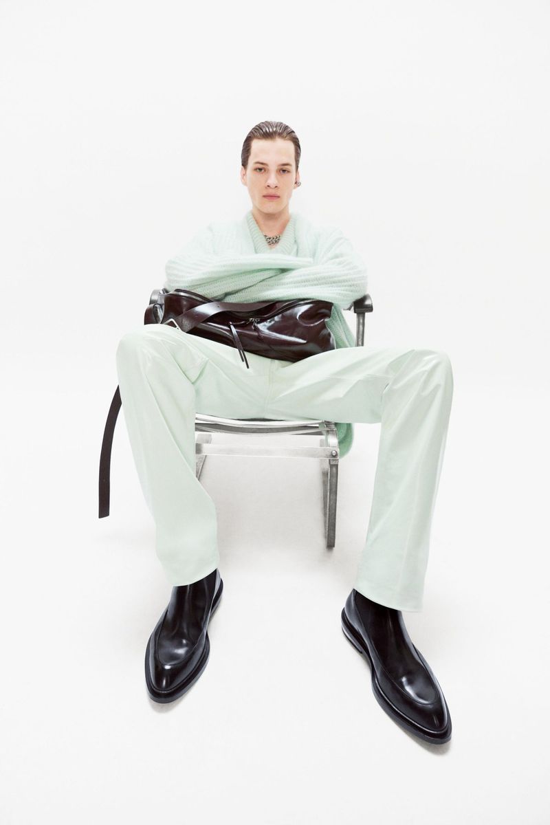 Stylish adult male seated in a clear chair against a bright white backdrop, wearing a light green pajama-style outfit and shiny black dress shoes.