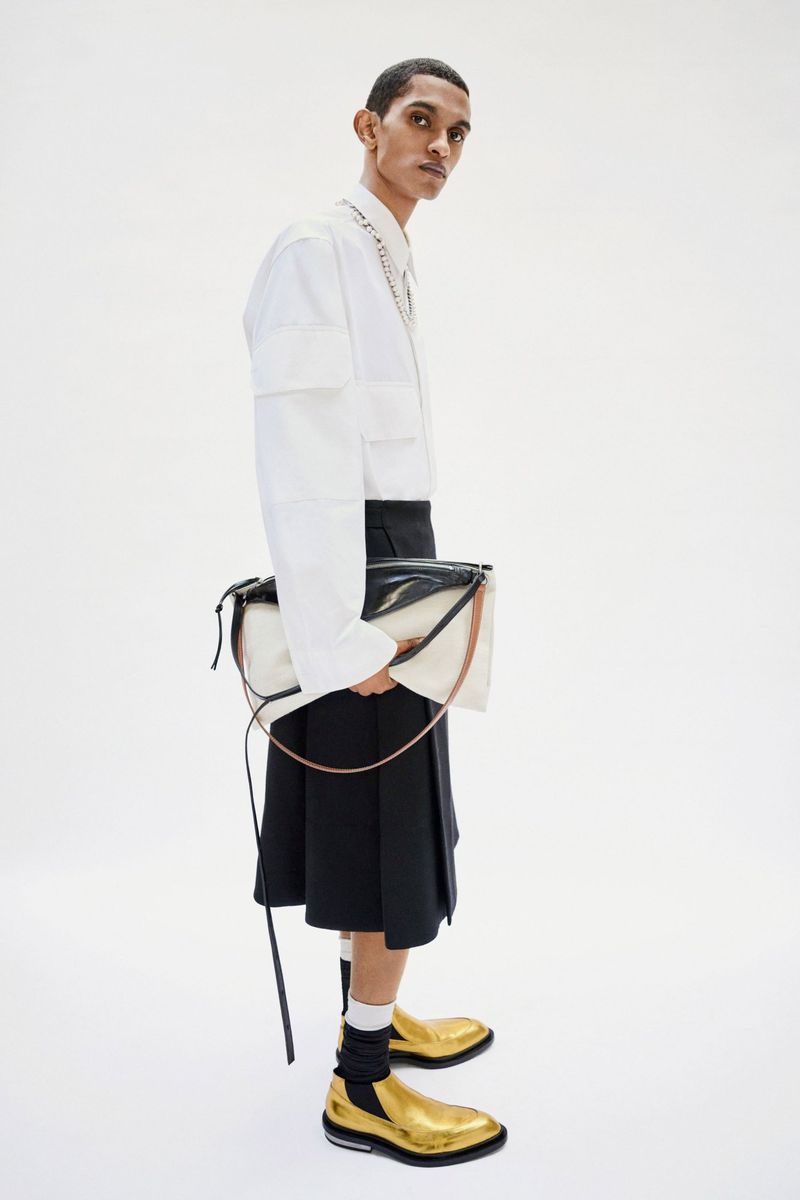 Fashionable adult male poses in a white shirt, black kilt, gold shoes, and a black-and-white purse against a plain white backdrop.