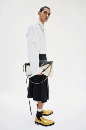 Fashionable adult male poses in a white shirt, black kilt, gold shoes, and a black-and-white purse against a plain white backdrop.