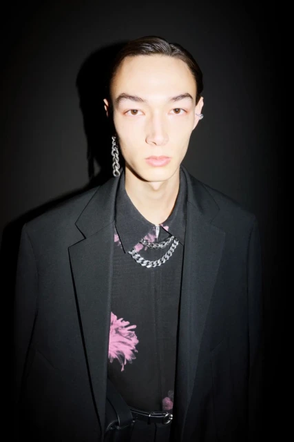 Portrait of a stylish adult male with slicked-back hair wearing a black suit jacket over a floral shirt and chain necklaces.