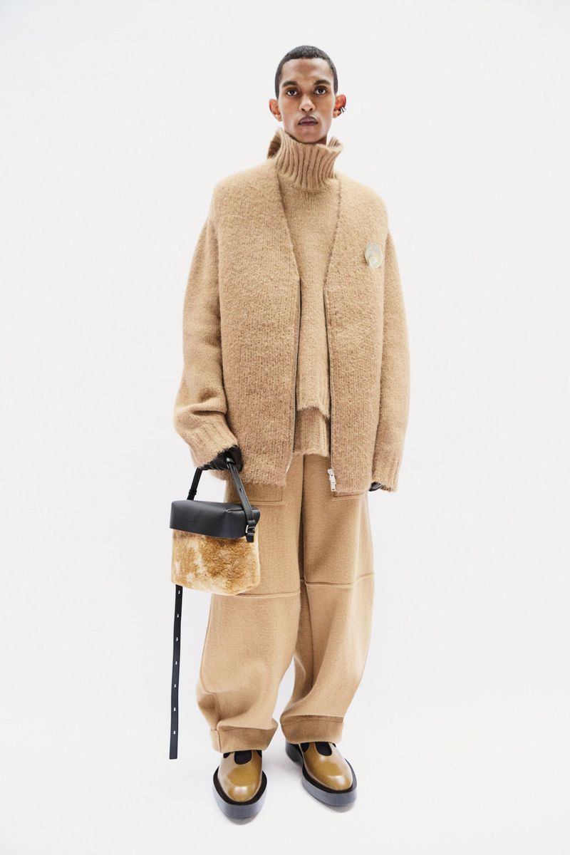 Adult male fashion model poses in a tan wool knit ensemble with handbag and patent leather shoes against a white background.