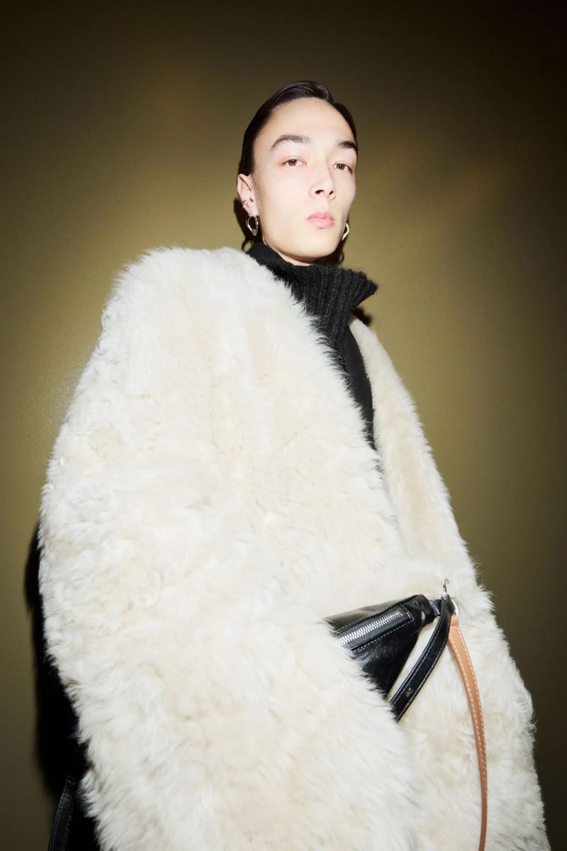 Portrait of a young adult in a black turtleneck and a large, fluffy white coat, posing in front of a dark olive green background.
