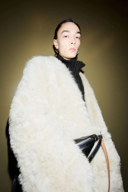 Portrait of a young adult in a black turtleneck and a large, fluffy white coat, posing in front of a dark olive green background.