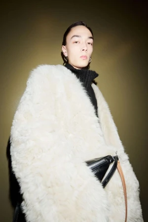Portrait of a young adult in a black turtleneck and a large, fluffy white coat, posing in front of a dark olive green background.