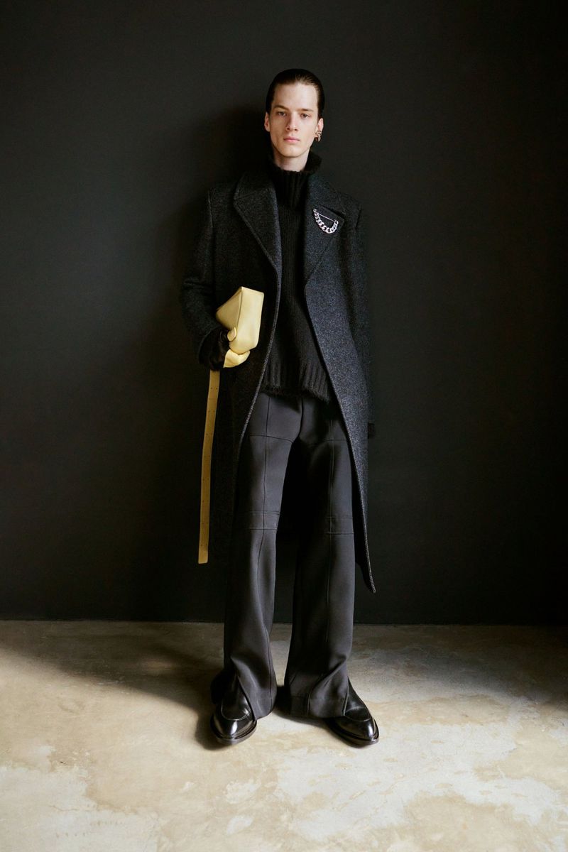 A young man wearing an overcoat and carrying a clutch stands in a dark grey studio with visible floor texturing. Fashion editorial.
