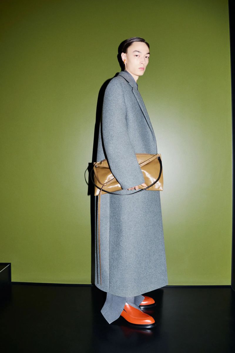 Fashion shot of a model wearing a long grey wool coat and orange leather loafers, holding a tan leather purse against a green wall.