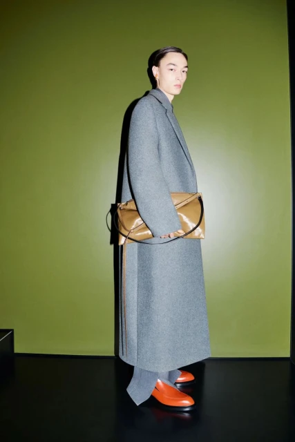 Fashion shot of a model wearing a long grey wool coat and orange leather loafers, holding a tan leather purse against a green wall.