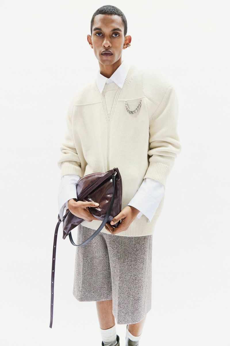 Male model poses in an off-white sweater, gray shorts, and black leather bag against a bright, seamless studio backdrop.