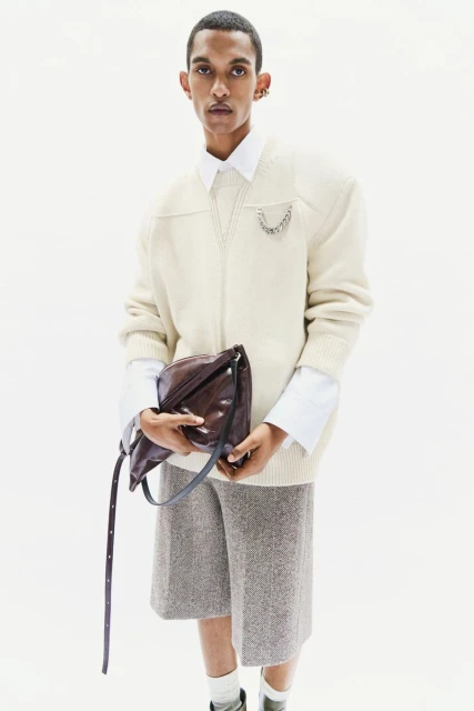 Male model poses in an off-white sweater, gray shorts, and black leather bag against a bright, seamless studio backdrop.