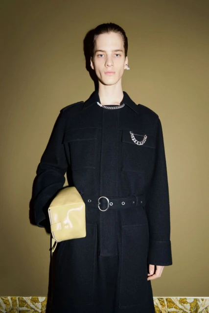 A fair-skinned young adult in a black jacket is holding a small purse in a studio against a textured brown wall with flooring detail.