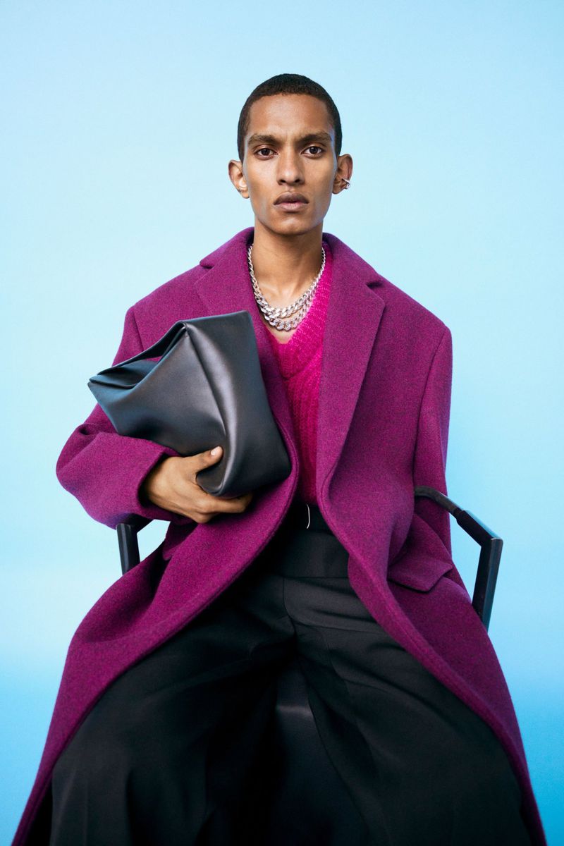 Male model in magenta coat and black pants holds structured black bag, set against a bright blue studio backdrop in a fashion shot.