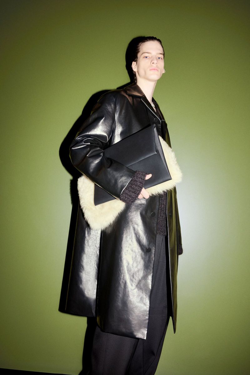 Adult male fashion model poses in a shiny black leather coat with fur trim, holding a black leather clutch against a green background.
