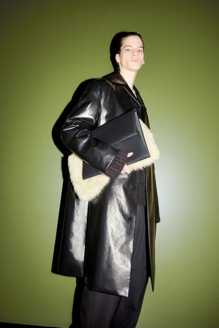Adult male fashion model poses in a shiny black leather coat with fur trim, holding a black leather clutch against a green background.
