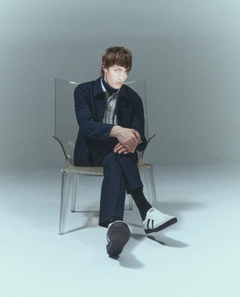 Stylish man in a blue jacket and black turtleneck poses seated on a transparent chair in a bright studio setting, looking at camera.