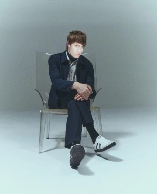 Stylish man in a blue jacket and black turtleneck poses seated on a transparent chair in a bright studio setting, looking at camera.