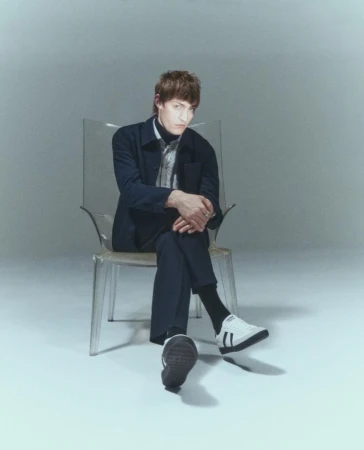 Stylish man in a blue jacket and black turtleneck poses seated on a transparent chair in a bright studio setting, looking at camera.