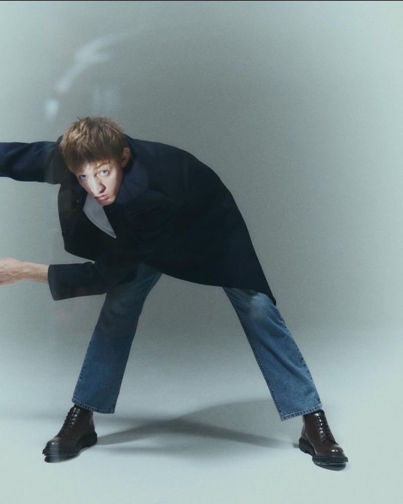 A young adult bends forward in a navy coat, blue jeans, and dark brown boots in a simple studio environment with natural lighting.
