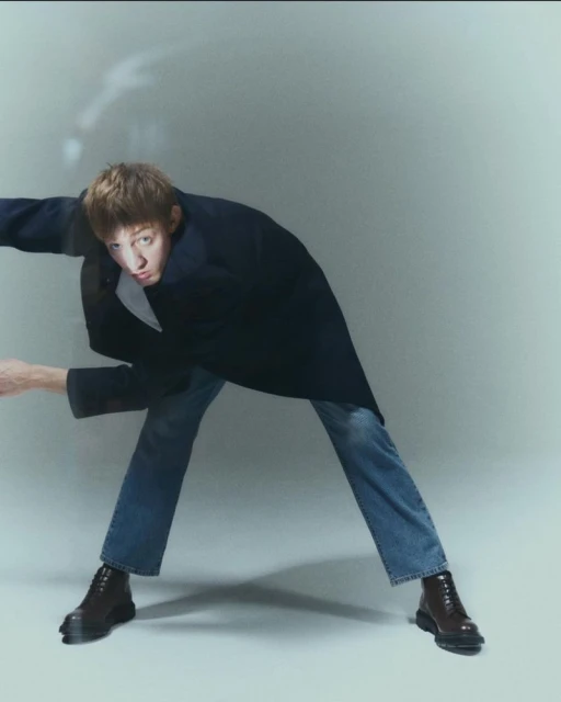 A young adult bends forward in a navy coat, blue jeans, and dark brown boots in a simple studio environment with natural lighting.