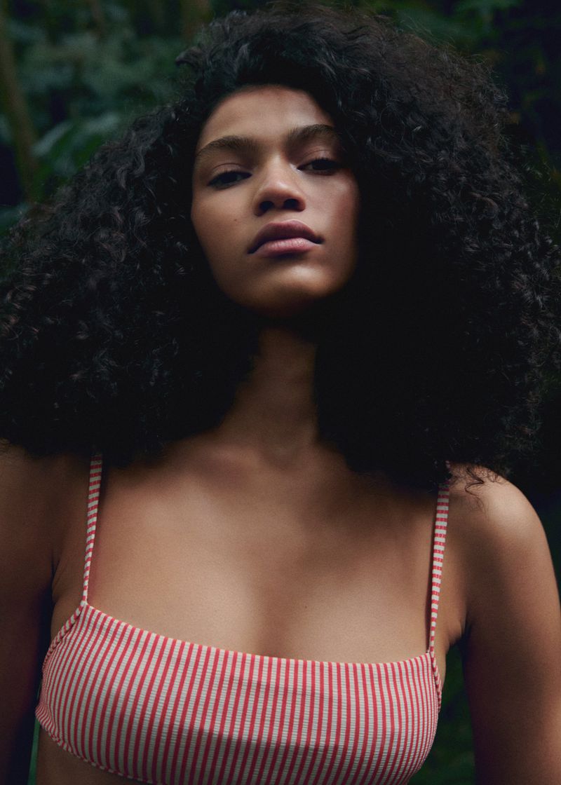 Portrait of a woman with dark curly hair wearing a red and white striped bikini top against a backdrop of dark green foliage.