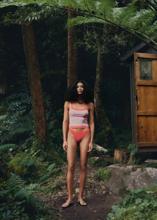 A woman poses in a pink top and coral-colored bottoms amidst a lush forest, next to a small wooden outhouse. Natural light.
