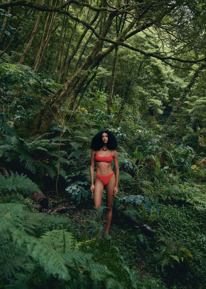 A woman in a red swimsuit standing in a lush green forest, surrounded by ferns and trees, bathed in diffused natural light.