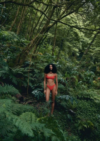 A woman in a red swimsuit standing in a lush green forest, surrounded by ferns and trees, bathed in diffused natural light.