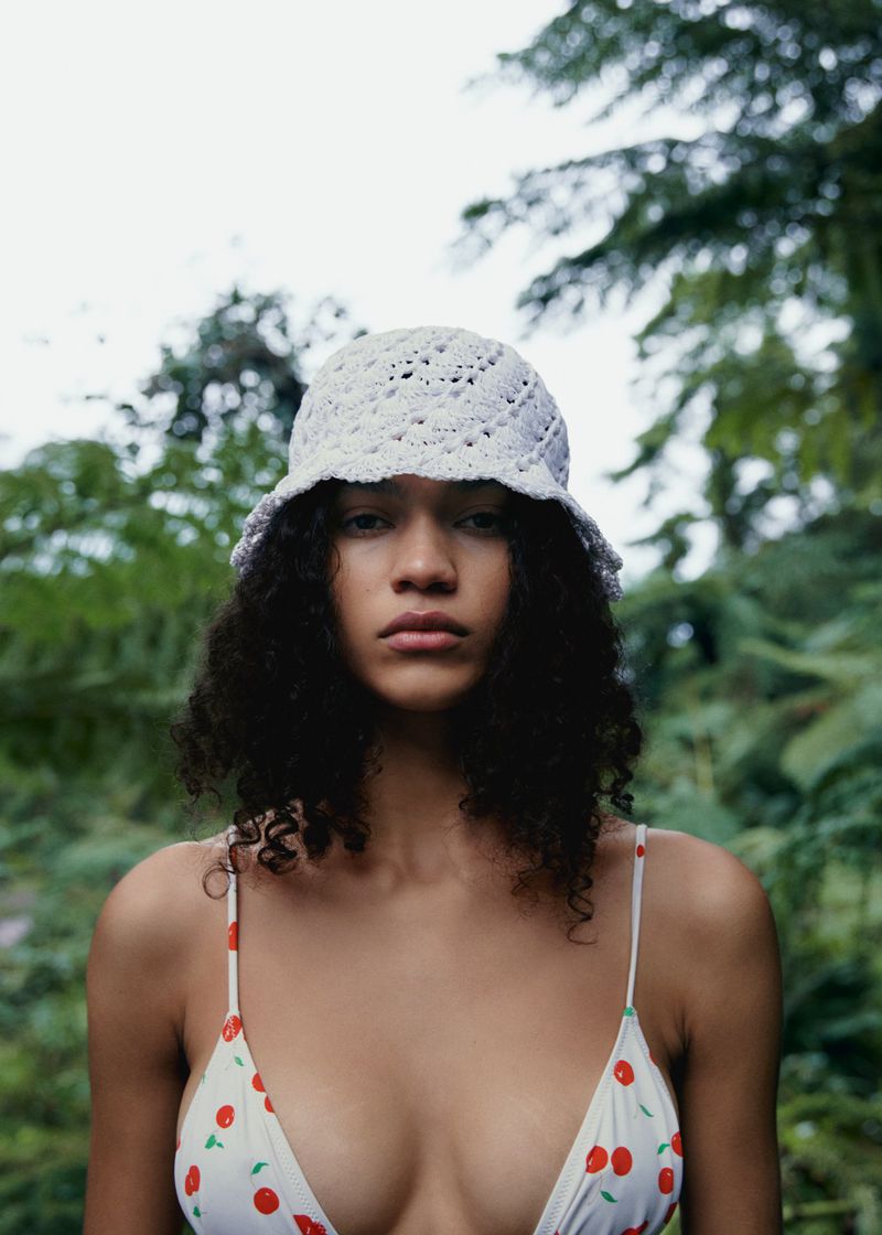 Woman in a cherry-print top and crocheted hat poses outdoors with a serious expression, surrounded by trees and greenery.