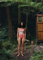 Raynara Negrine in a pink tank top and coral bikini bottoms standing on a dirt forest path beside a rustic wooden shed.