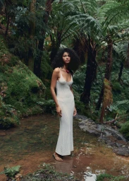 Raynara Negrine in a white knit maxi dress standing in a forest stream surrounded by lush green ferns.