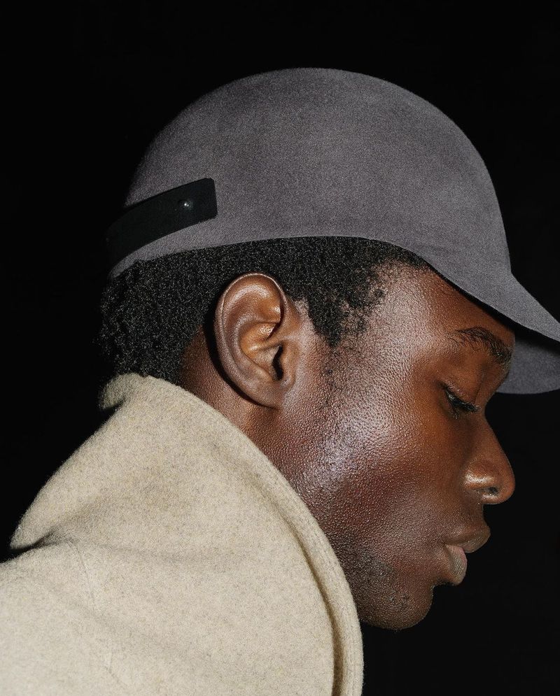 Portrait of a black man wearing a gray cap and tan overcoat, shot in a dark indoor environment with shallow depth of field.