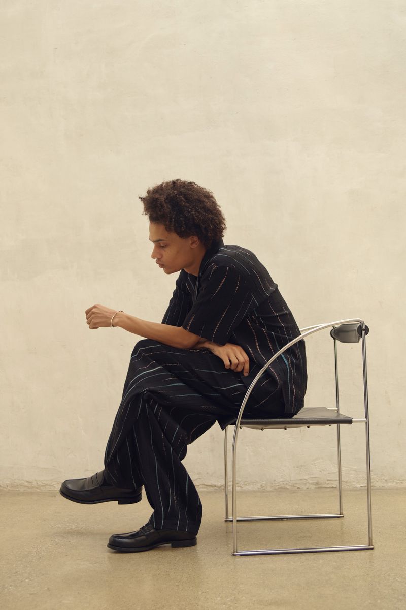 Person in black pinstripe suit sitting on metal and leather chair in a minimalist studio against a neutral wall and floor.