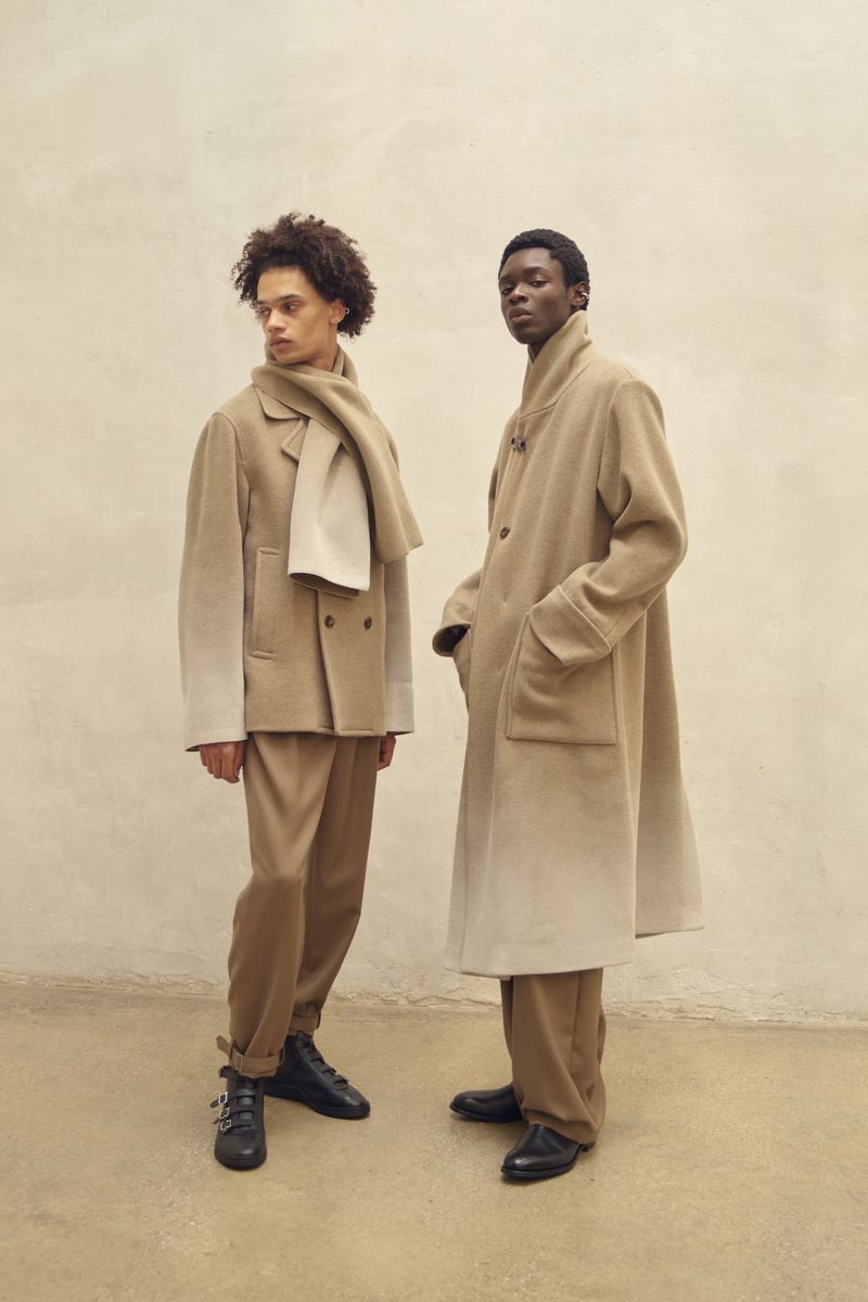 Two dark-skinned male fashion models in beige coats and pants stand in front of a light beige wall in a studio setting.