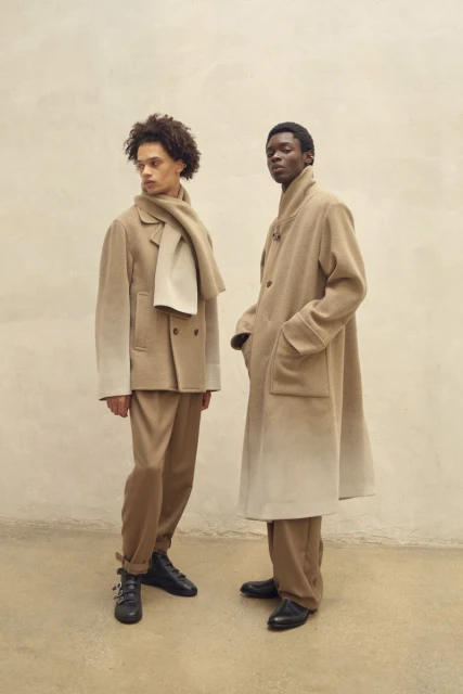 Two dark-skinned male fashion models in beige coats and pants stand in front of a light beige wall in a studio setting.