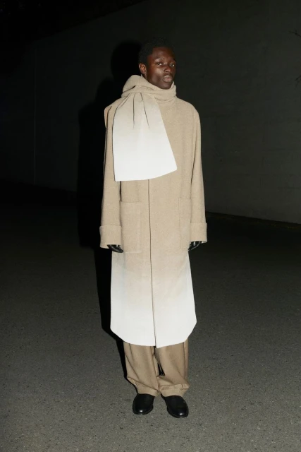 An adult male model poses in a beige coat and draped scarf in an outdoor setting, illuminated by dark, moody lighting.