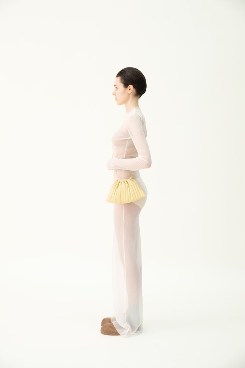 Woman in sheer white outfit poses with a yellow woven purse in a studio setting against a stark white backdrop, fashionably minimalist.