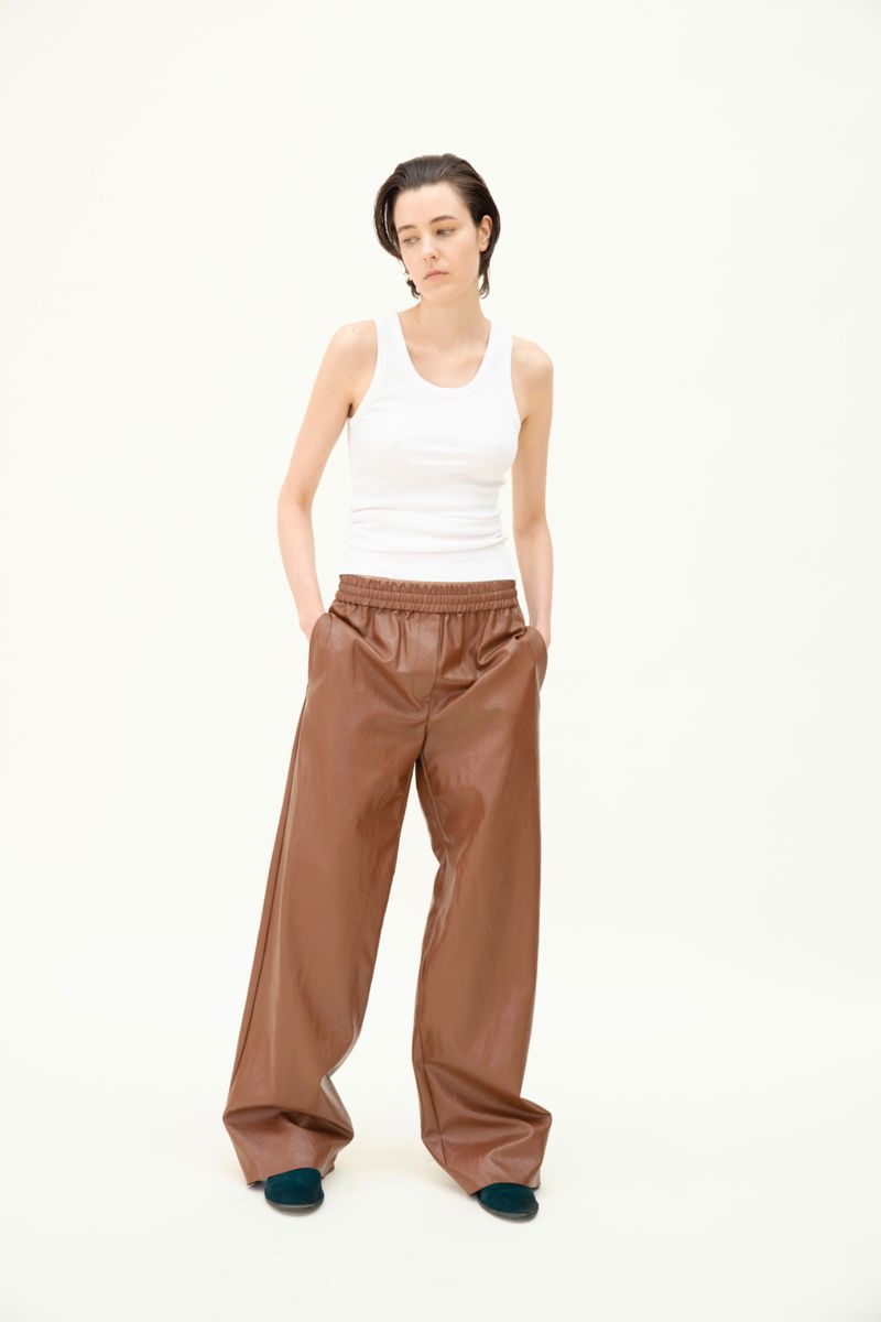 Adult woman in a white tank top and brown leather pants poses in a photo studio setting with a white backdrop and neutral lighting.