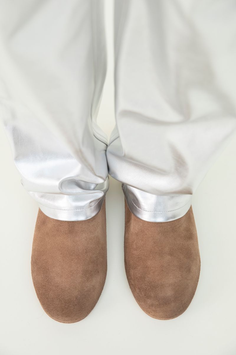 A pair of brown suede clogs are visible below silver satin pants, set against a white background in a studio setting.