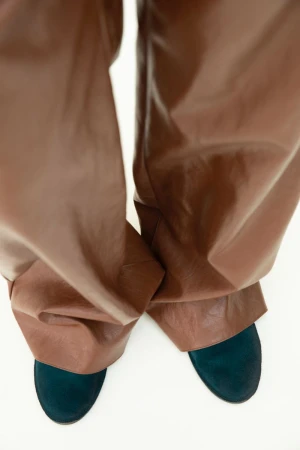 Eye-level shot showcasing brown leather pants paired with blue suede shoes, set against a stark white background.