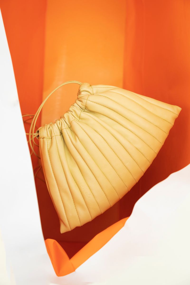 Pale yellow, vertically pleated purse with drawstrings displayed against a bright orange and white backdrop, product photography.