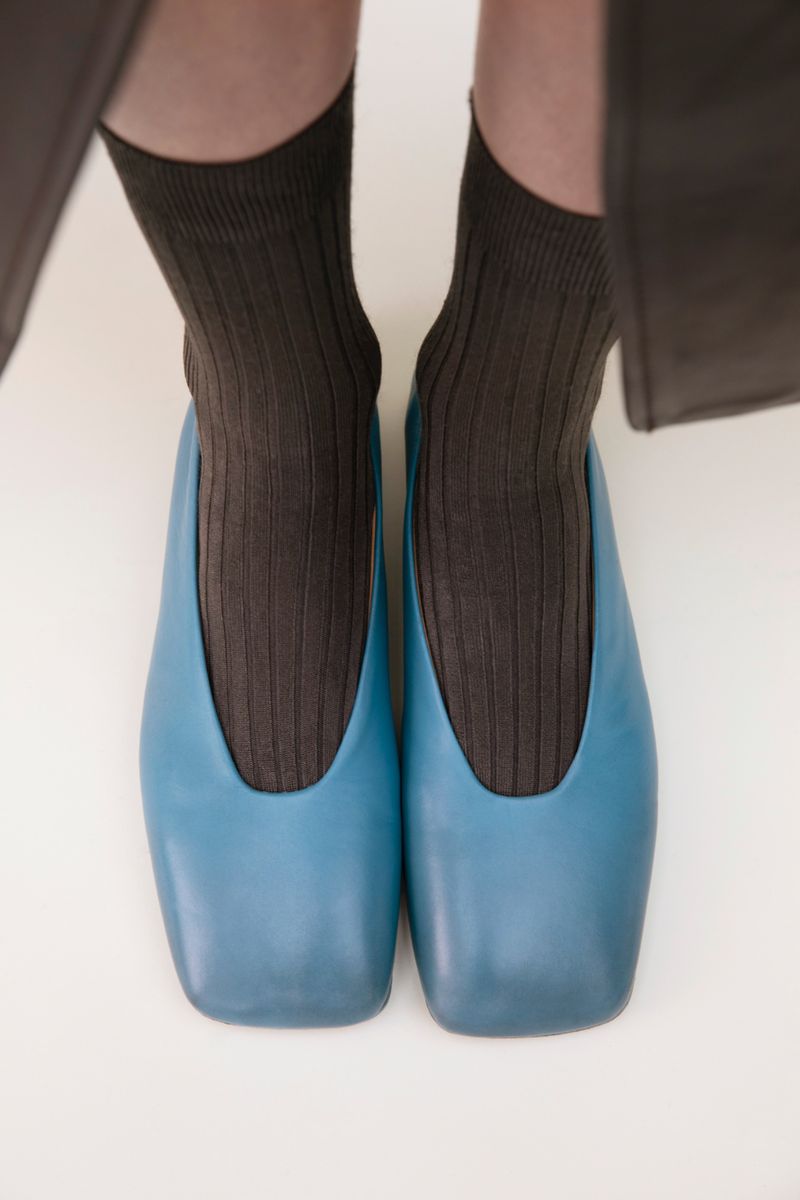 A close-up of blue leather ballet flats worn with ribbed brown socks, set against a plain white background.