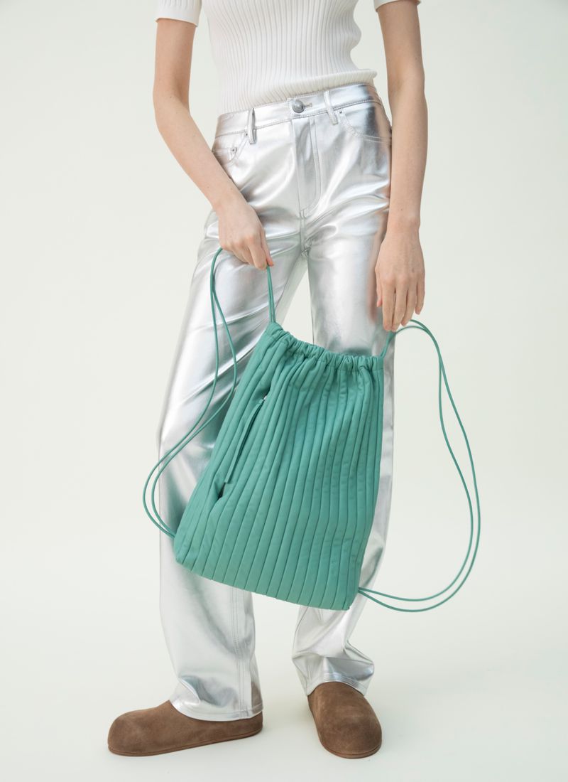 Studio shot of person wearing metallic pants holding a green knitted string backpack with brown footwear on a white backdrop.