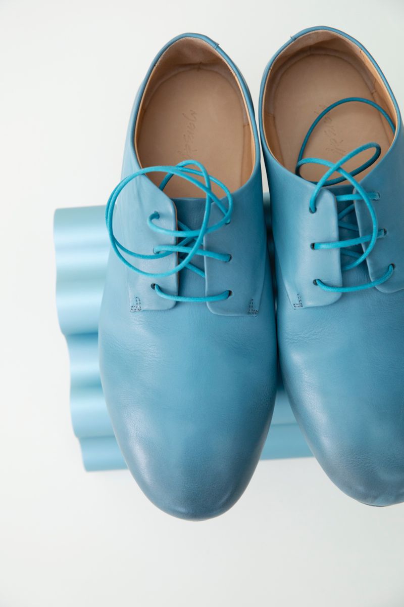 Pair of light blue leather dress shoes with turquoise laces displayed on a blue, corrugated stand, set against a white background.