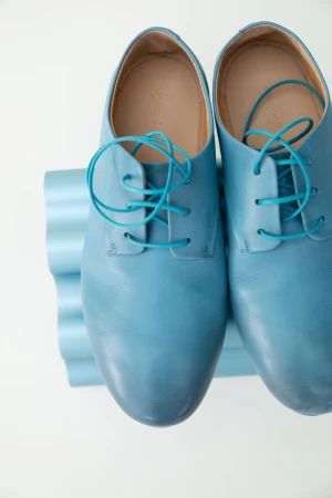 Pair of light blue leather dress shoes with turquoise laces displayed on a blue, corrugated stand, set against a white background.