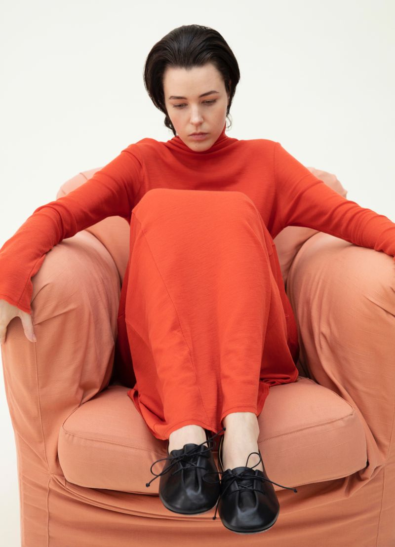 A woman in a red dress and black flats sits on an apricot-colored upholstered chair against a white background, looking down.