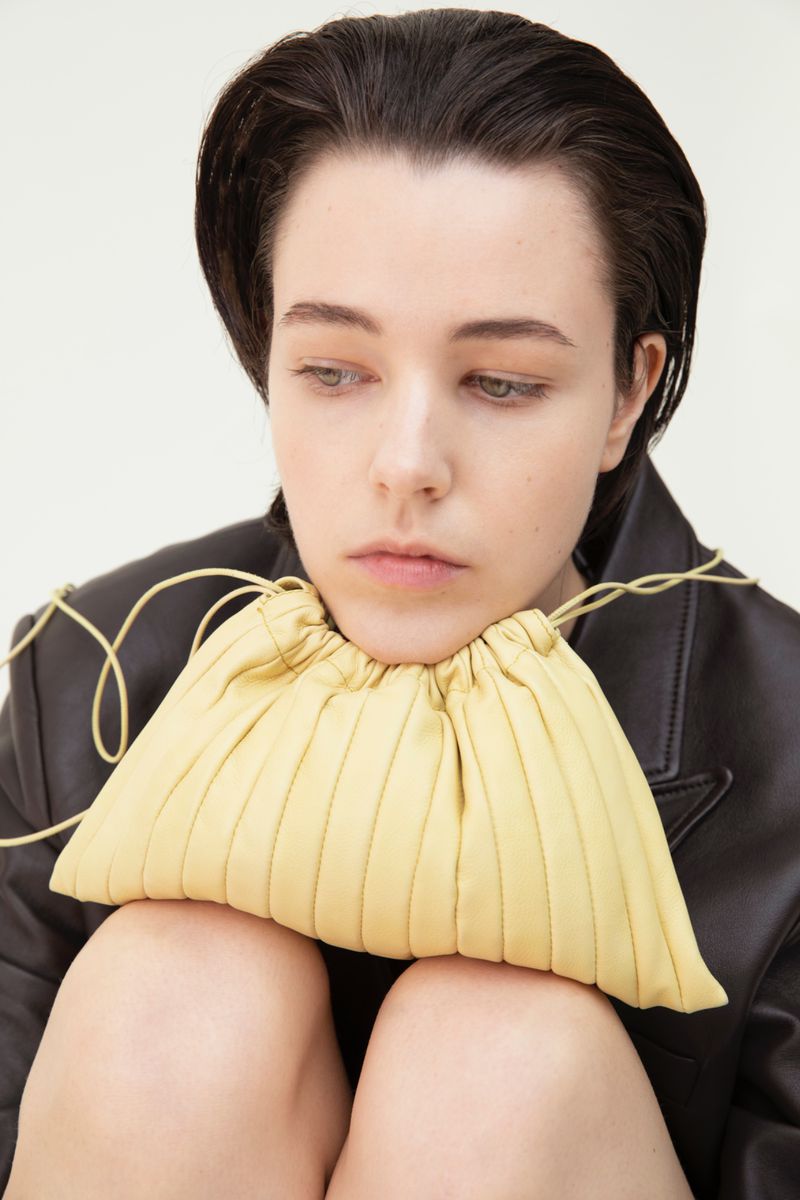 Model in a black leather blazer resting her chin on a pale yellow pleated drawstring clutch bag against a white studio background.