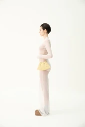 Model standing in profile wearing a sheer white long-sleeved dress and holding a pale yellow pleated pouch against a white background.