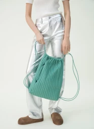 Model wearing metallic silver trousers and a white top holding a green quilted drawstring bag against a white studio background.